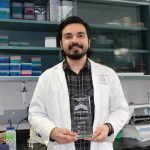 Student wearing a lab coat holds a glass trophy with 3MT logo on it.