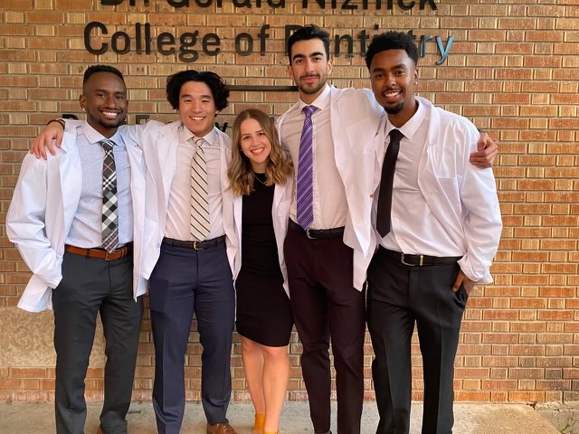 A group of dentistry students stand wearing white coats, smiling