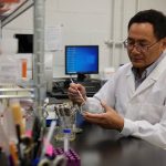 Dr. Kangmin Duan, wearing a white lab coat, sits in his lab. He is holding an instrument and petri dish.