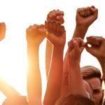 People pump their fists in the air in front of a bright sun.