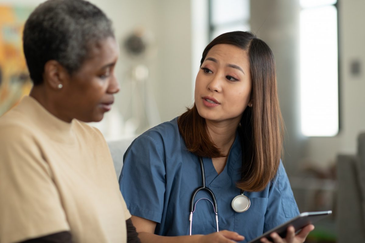 A young health-care professional of Asian descent and an elderly patient of African discuss the patient’s care while looking at a tablet computer.