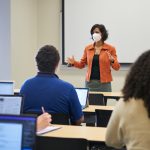 Instructor in KN95 mask at the front of a classroom full of students with backs to camera