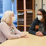 Two people wearing masks have a conversation while sitting at a table.