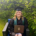 Woman sitting in patio chair, outdoors in front of green trees. She is wearing a grad cap and gown and holding her degree.