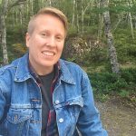Fenton Litwiller, wearing a jean jacket, smiles in the woods