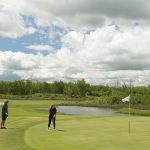 Two golfers standing on the green getting ready to putt. The sky is bright with broken cloud.