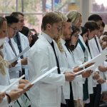 Students wearing white coats each hold a piece of paper. They are reading the Physician's Pledge.