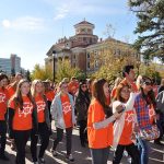 A crowd of students, faculty and university staff walk past the University of Manitoba administration building wearing orange shirts.