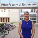Dr. Marcia Friesen standing, smiling and posting for the camera in front of the E I T C - S P C sky walk. On the silver skywalk in black letters it reads "Price Faculty of Engineering. Marcia is wearing a blue dress. She is outside on a sunny day with a full bike rack in the background.