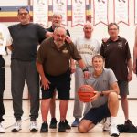 Eight men grouped together in gymnasium, smiling.