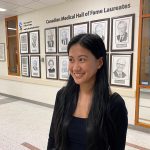 Valcourt poses for photo in front of 12 framed drawings of the CMHF laureates. Text on the wall reads "Canadian Medical Hall of Fame Laureates."