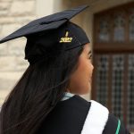 A UM grad in cap and down looks off into the distance. Their tassle has 2022 on it and they stand in front of a limestone brick building with stained glass doors.