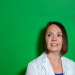Biochemistry and medical genetics Prof. Tamra Werbowetski-Ogilvie stands in a labcoat in front of a plain green background, looking up.
