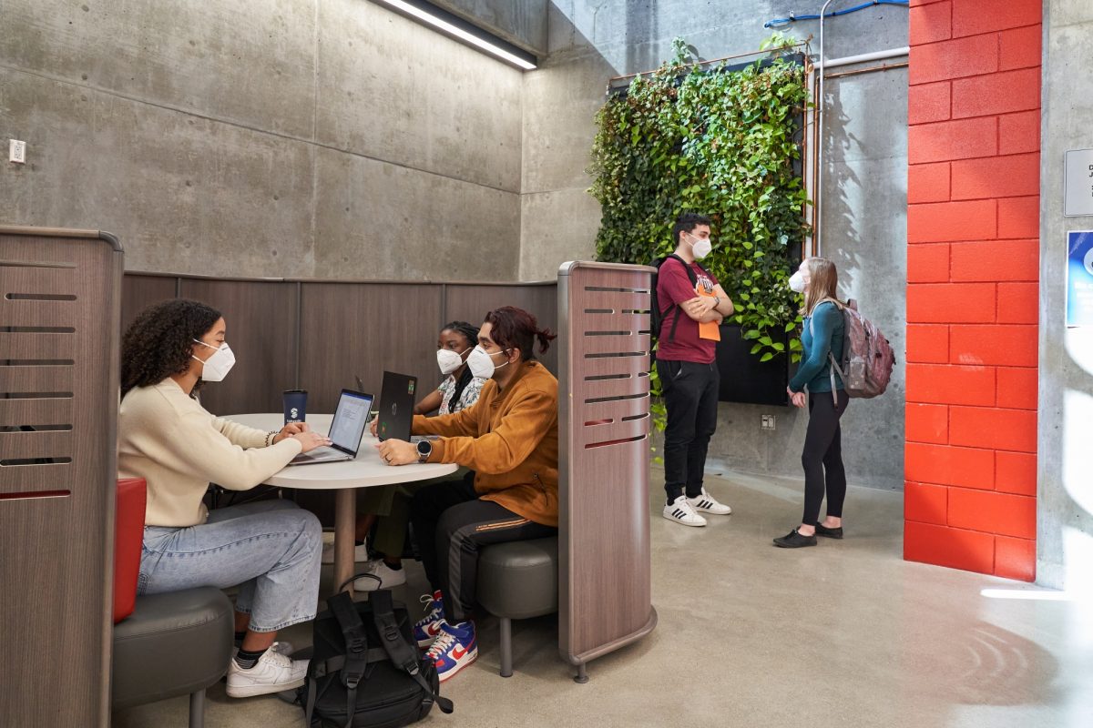 Three students study together in a booth. Two other students stand chatting in the background. They are all wearing KN-95 masks.