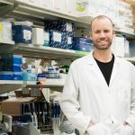 Brent Guppy stands in his lab wearing a white lab coat.