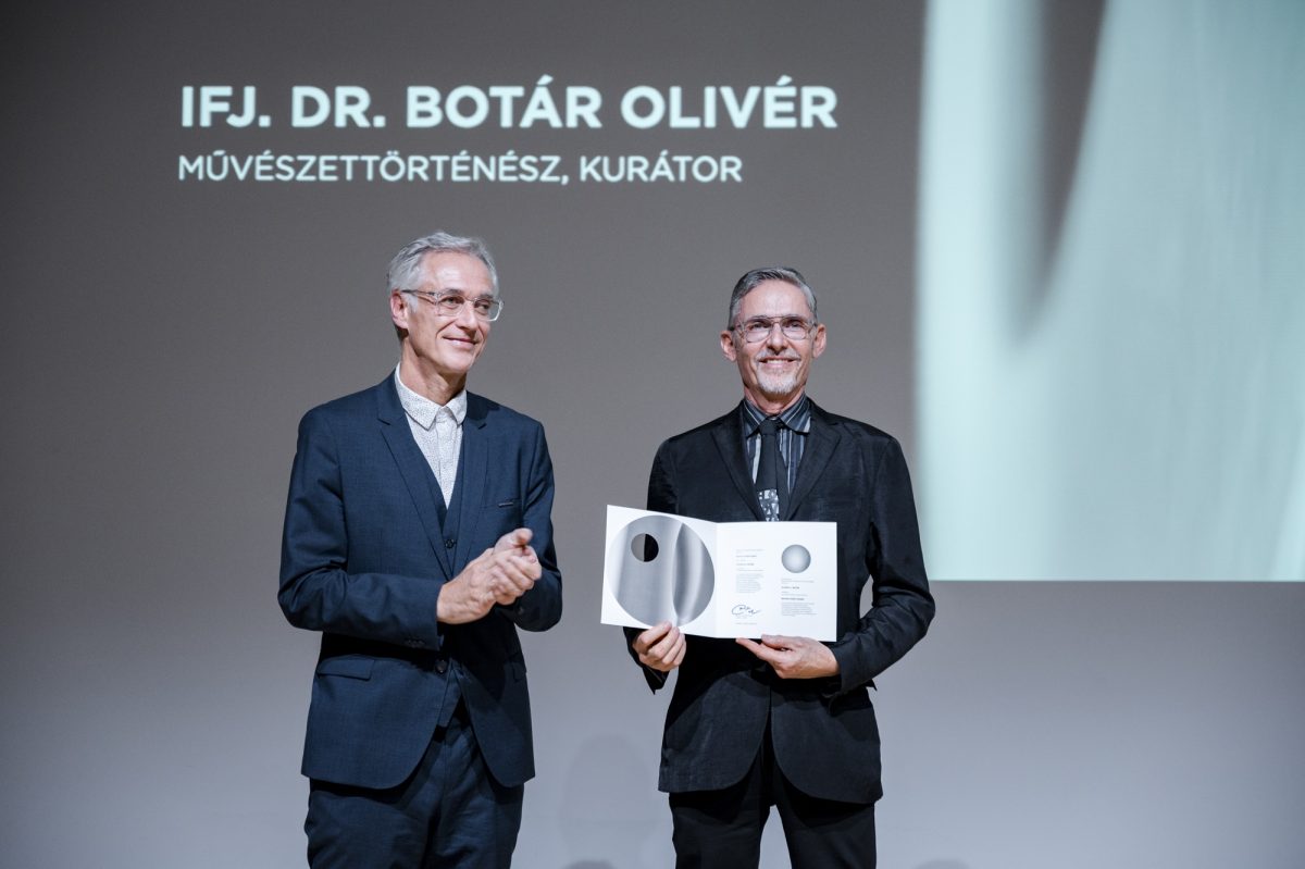 Dr. Ovliver Botar stands on stage with Rector József Fülöp and holds a glass award