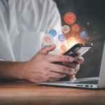 Person at a computer holding a phone with social media icons popping out.