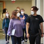 Two students featured walking together indoors, while masked., various other students, also masked are walking by.