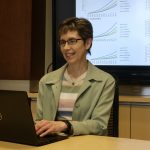 Dr. Lisa Lix sits at a table and uses a laptop. On a screen behind her are six graphs.