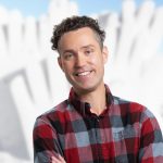 Man in plaid shirt with arms crossed, a snow sculpture behind him.