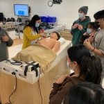 Students wearing stethoscopes gather around instructor. One person places stethoscope on chest of training manikin.