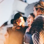 Three women stand outside a building in the warm sun. They are smiling and laughing as they talk with each other.