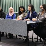 The physicians and students sit at a table. Two microphones sit on the table.
