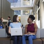 Two smiling people sit on a bench chatting with each other. One has a laptop in her lap while the other has a notebook.
