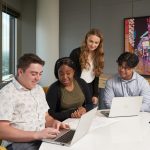 Four students looking at two computers with a big window in the background.