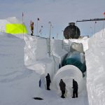 A large snow trench being built. Three people are visible in the trench, along with three giant balloons. A crane and bright yellow tent sit above the trench.