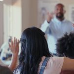 Students in class with their hands raised and an instructor at the front of the class.