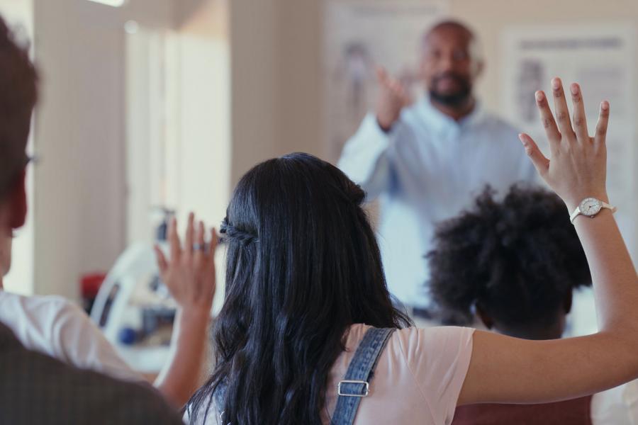 Students in class with their hands raised and an instructor at the front of the class.