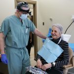 Catherine sits in a dental chair and wears a bib. Dr. McCoy stands next to her and wears scrubs, gloves and a face mask.