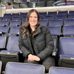 Woman with black jacket smiling and sitting in the stands in an arena.