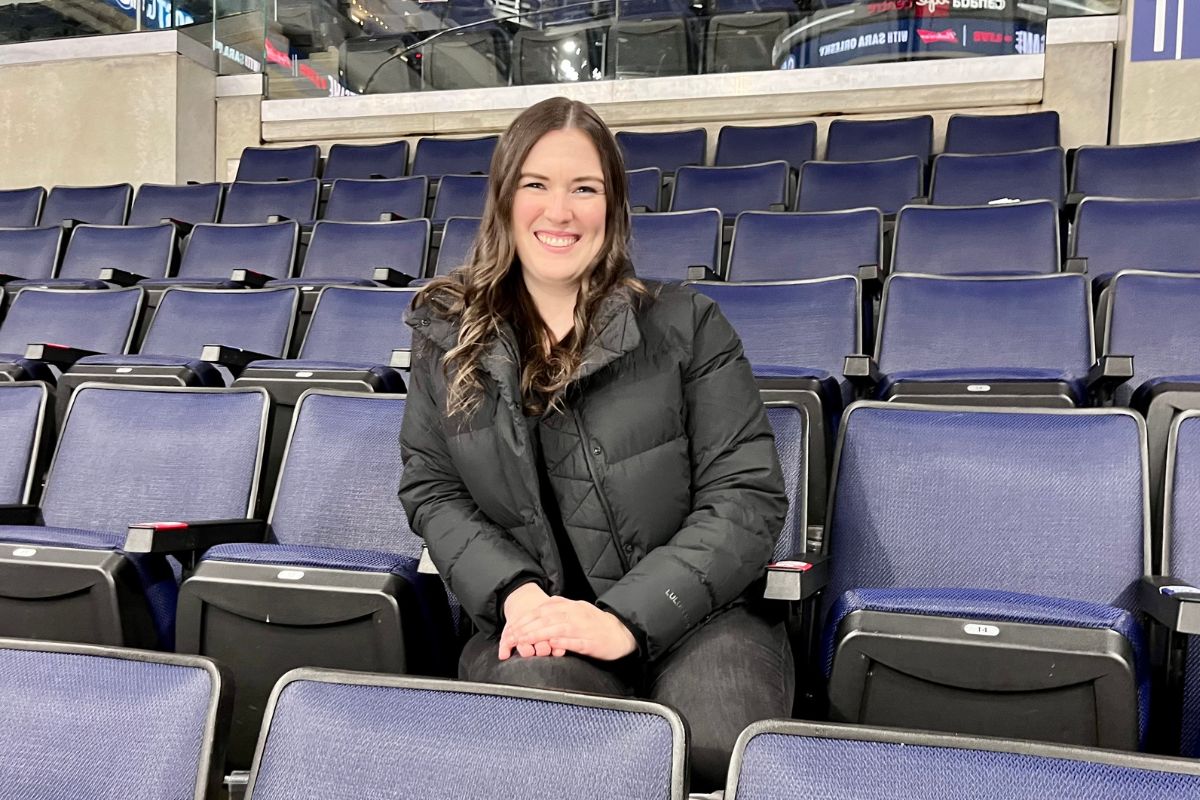 Woman with black jacket smiling and sitting in the stands in an arena.
