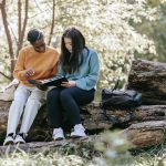 Two students sitting on a fallen tree in the forest doing school work