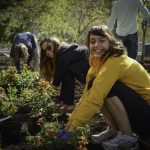 A woman in a yellow hoodie smiles at the camera and planting flowers.