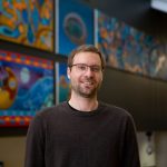 Man with short brown hair, close shaved brown beard wearing glasses, wearing a brown sweater.