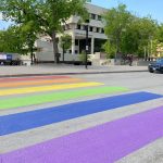 Crosswalk with purple, blue, green, yellow, orang and red stripes with green trees in the background.