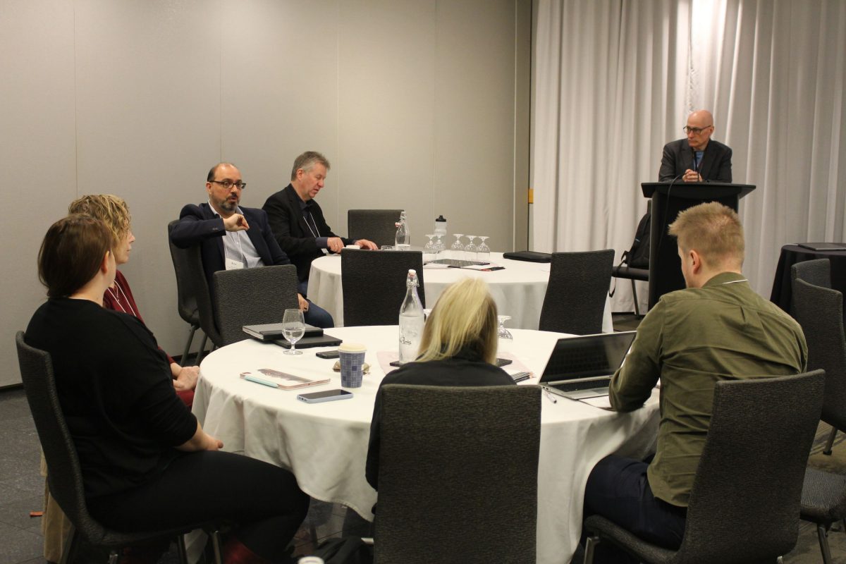 Four people sit at a round table. Drs. François and Jacobsen sit at another round table. Dr. Doupe stands at a lectern.