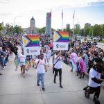 crowd of people walking in pride parade on sunny day, holding university of manitoba signs with pride colours