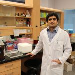 Portrait of Dr. Tanveer Sharif in his lab. He is wearing a lab coat. There is scientific equipment and bottles on the counter and on shelves behind him.