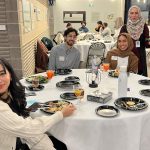 Three students sit at a table and another student is standing as they pose for a photo. Food is on their plates.