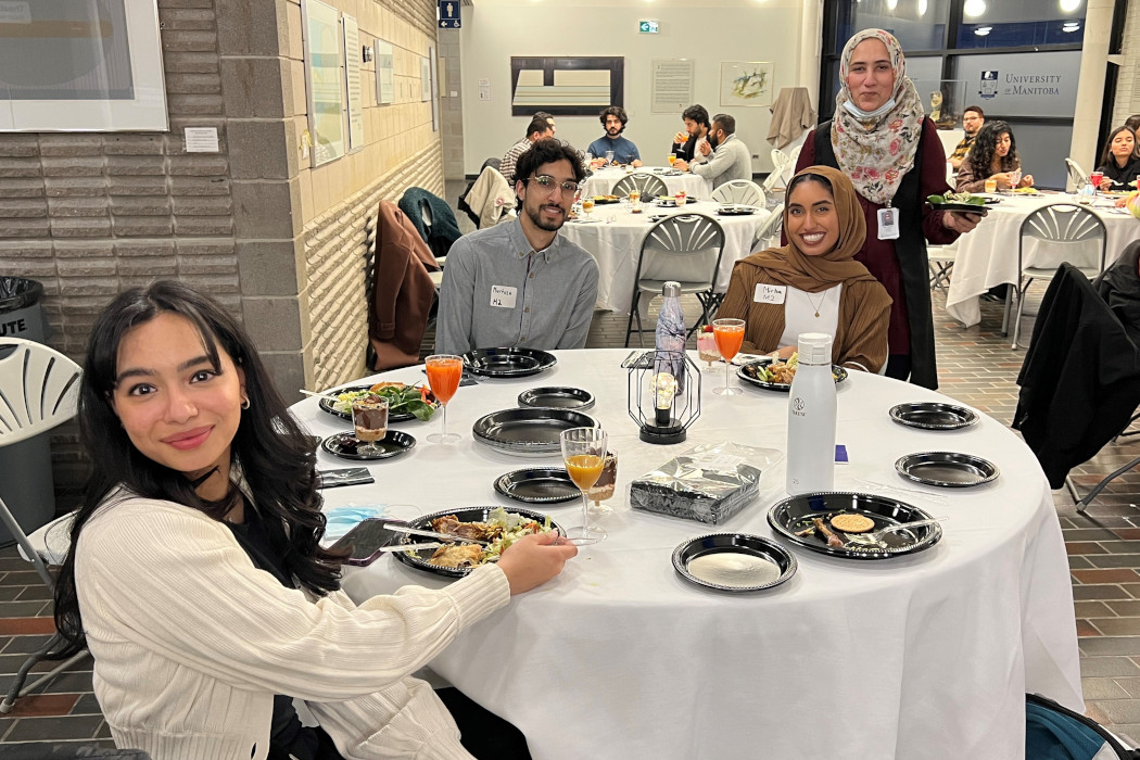 Three students sit at a table and another student is standing as they pose for a photo. Food is on their plates.