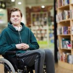 Content handsome young disabled student with headphones on neck siting in wheelchair and looking at camera in modern library or bookstore.