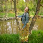 Female wearing a ribbon skirt and jean jacket standing in front of a river.