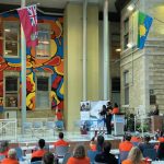 Mural featuring Indigenous symbols is unveiled. Blake Angeconeb stands near a lectern on the stage. People in the crowd are wearing orange shirts and some are taking photos of the mural.