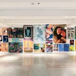 Dozens of vibrant, varying sizes painting hang on a white wall of the gallery.