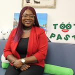 Dr. Olubukola-Olatosi sits on a small couch. Behind her is a sign that reads "tooth paste." The two "o's" in tooth form eyes and there are eyebrows above them and a mouth below. A smiling plush tooth with one tooth is also behind her.