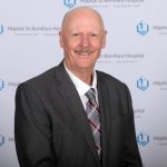 Portrait of Dr. Grant Pierce. In the background is the St. Boniface Hospital logo and it reads "Hopital St-Boniface Hospital, Recherche, Research," and "Foundation."